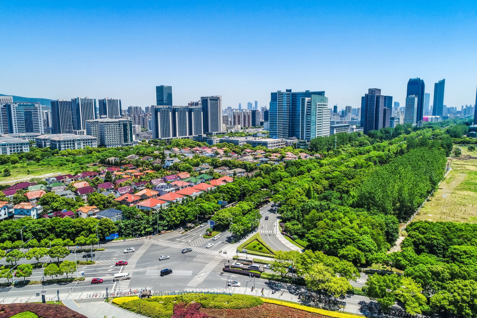 Aerial cityscape with green parkland, red-roofed buildings, and modern high-rises under a clear blue sky