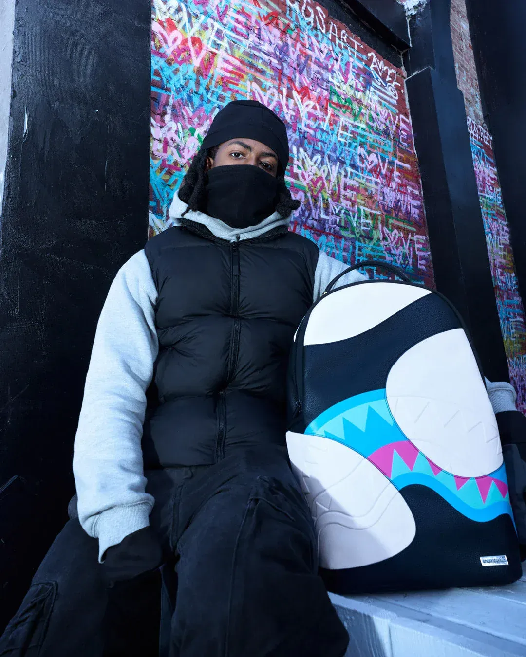 Persona mascherata con giacca imbottita nera e berretto, seduta accanto a un murale colorato raffigurante uno snowboard.