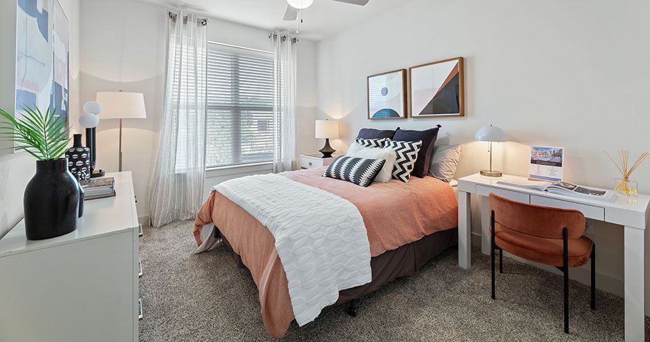 Bedroom with bed, desk, and dresser in neutral tones at The Standard at Domain NORTHSIDE, offers Domain apartments Austin. Orange accents, window with blinds.
