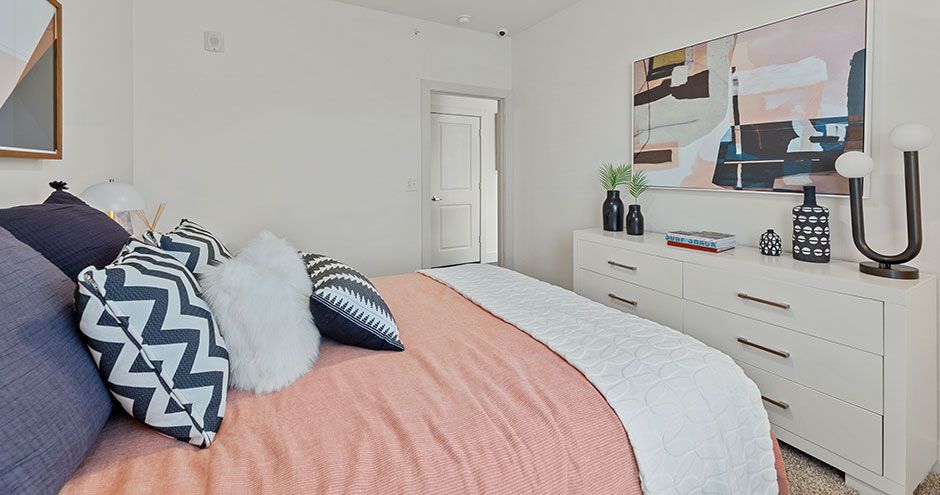 Bedroom with bed, pillows, white dresser, artwork, and a partially open door at The Standard at Domain NORTHSIDE, offers apartments at The Domain.