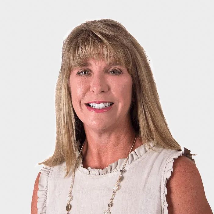 Blonde woman with bangs smiles, wearing a sleeveless light tan top and a necklace.