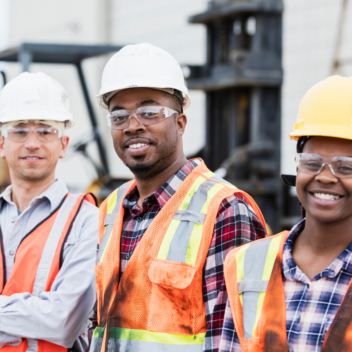 Three construction workers wearing hard hats, safety glasses, and orange vests smile at the camera.