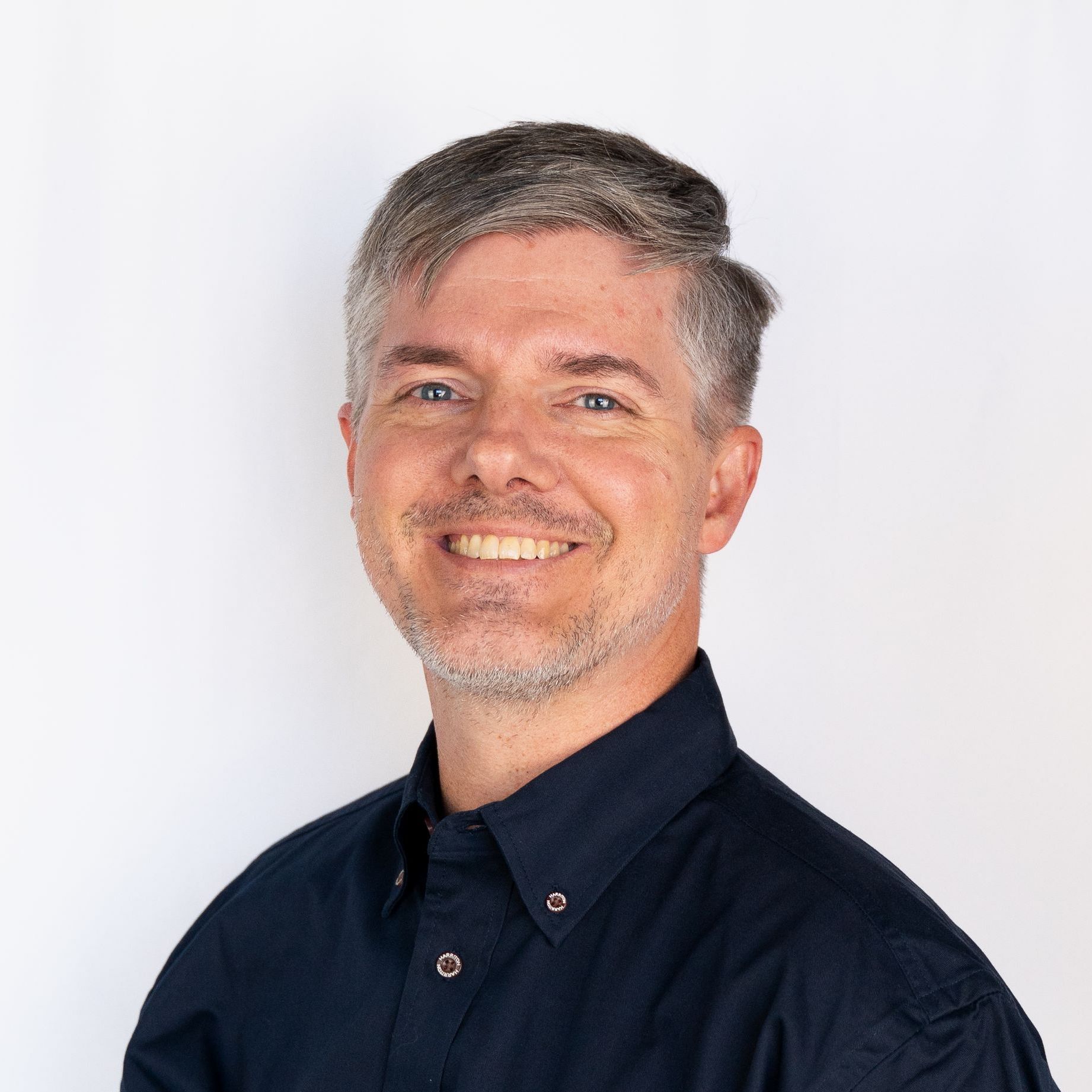 Man with graying hair and stubble smiles; he wears a dark blue button-down shirt.