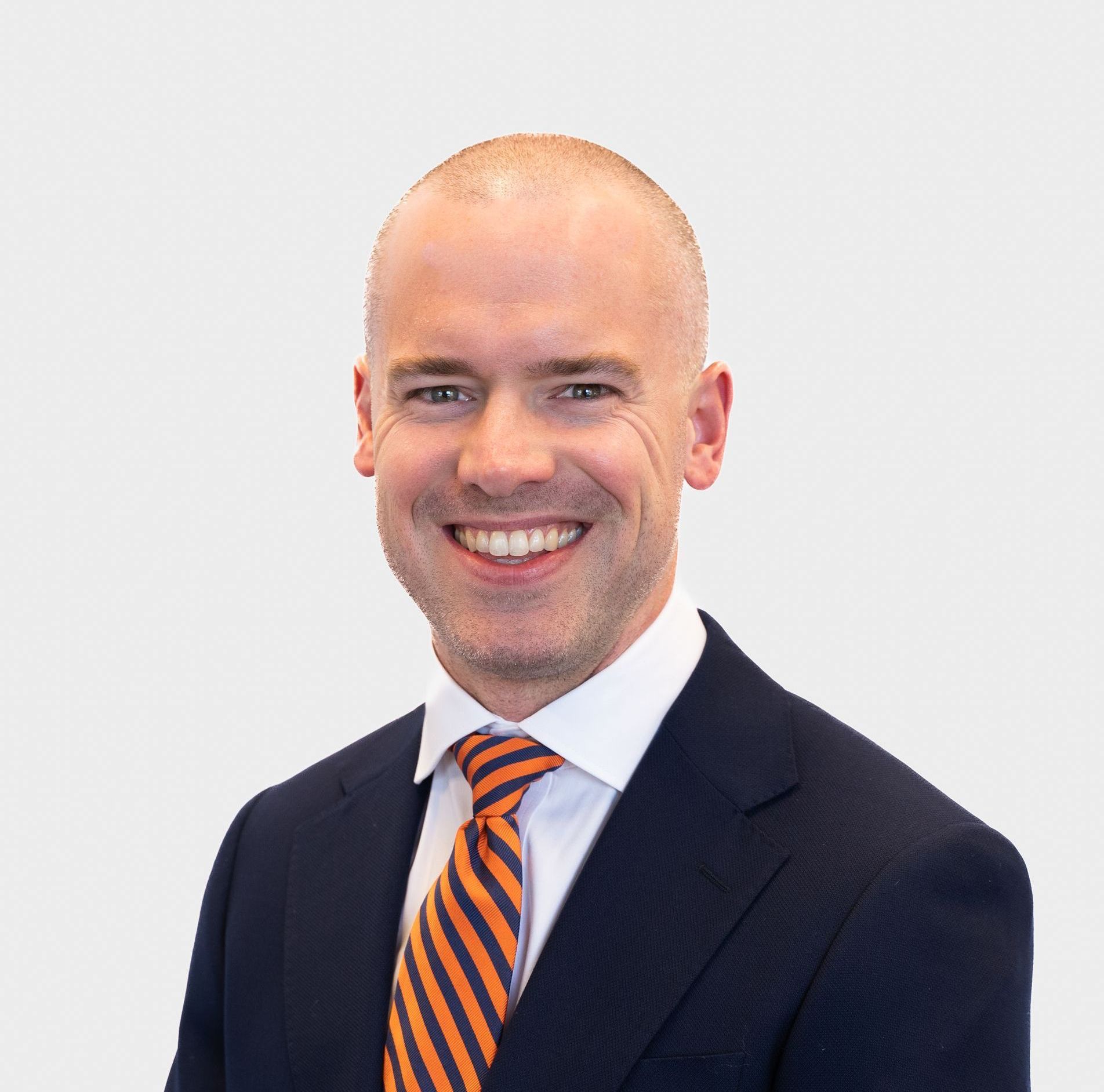 Man in a dark suit and orange striped tie smiles at the camera against a white backdrop.