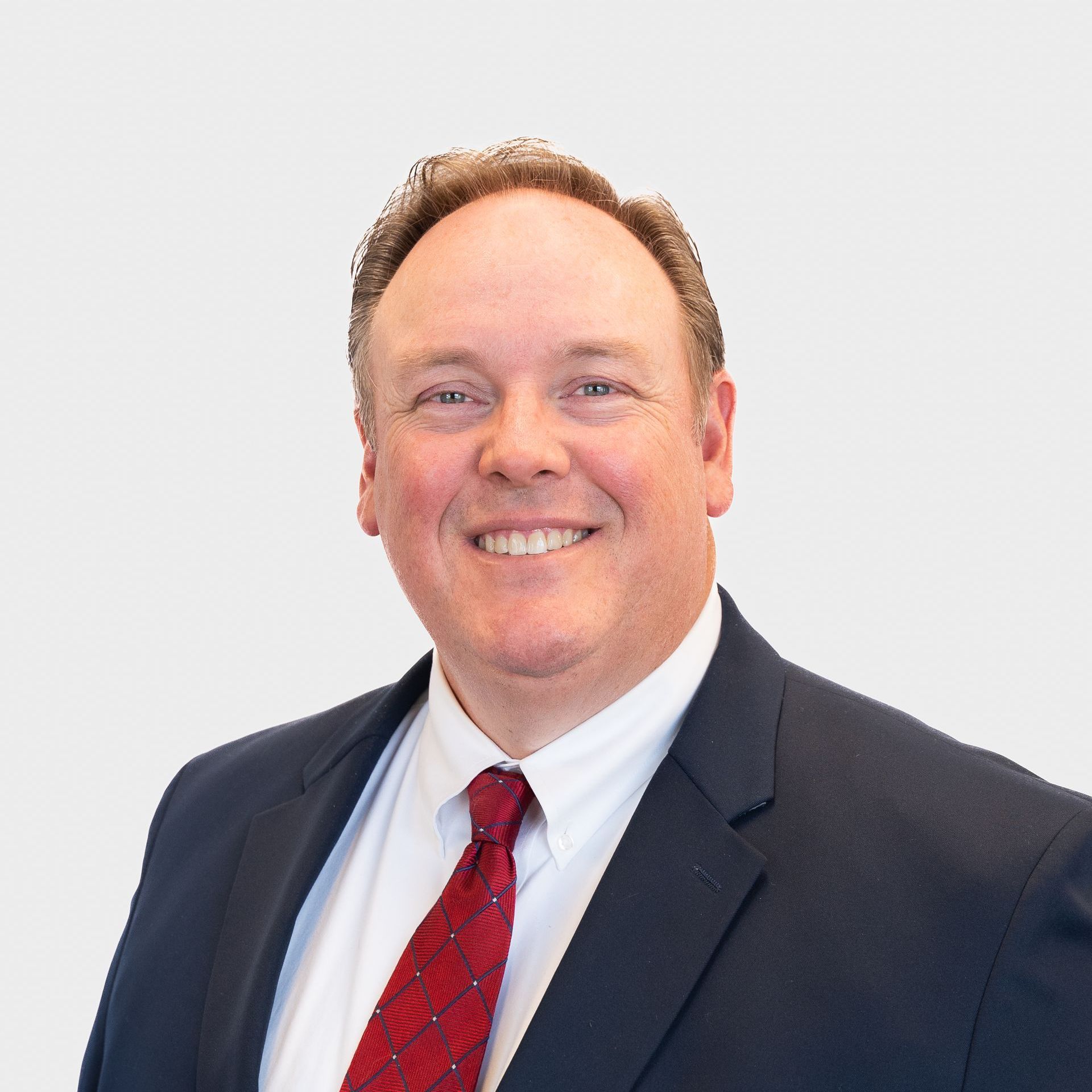 Man in a navy suit, white shirt, and red tie smiles against a white backdrop.
