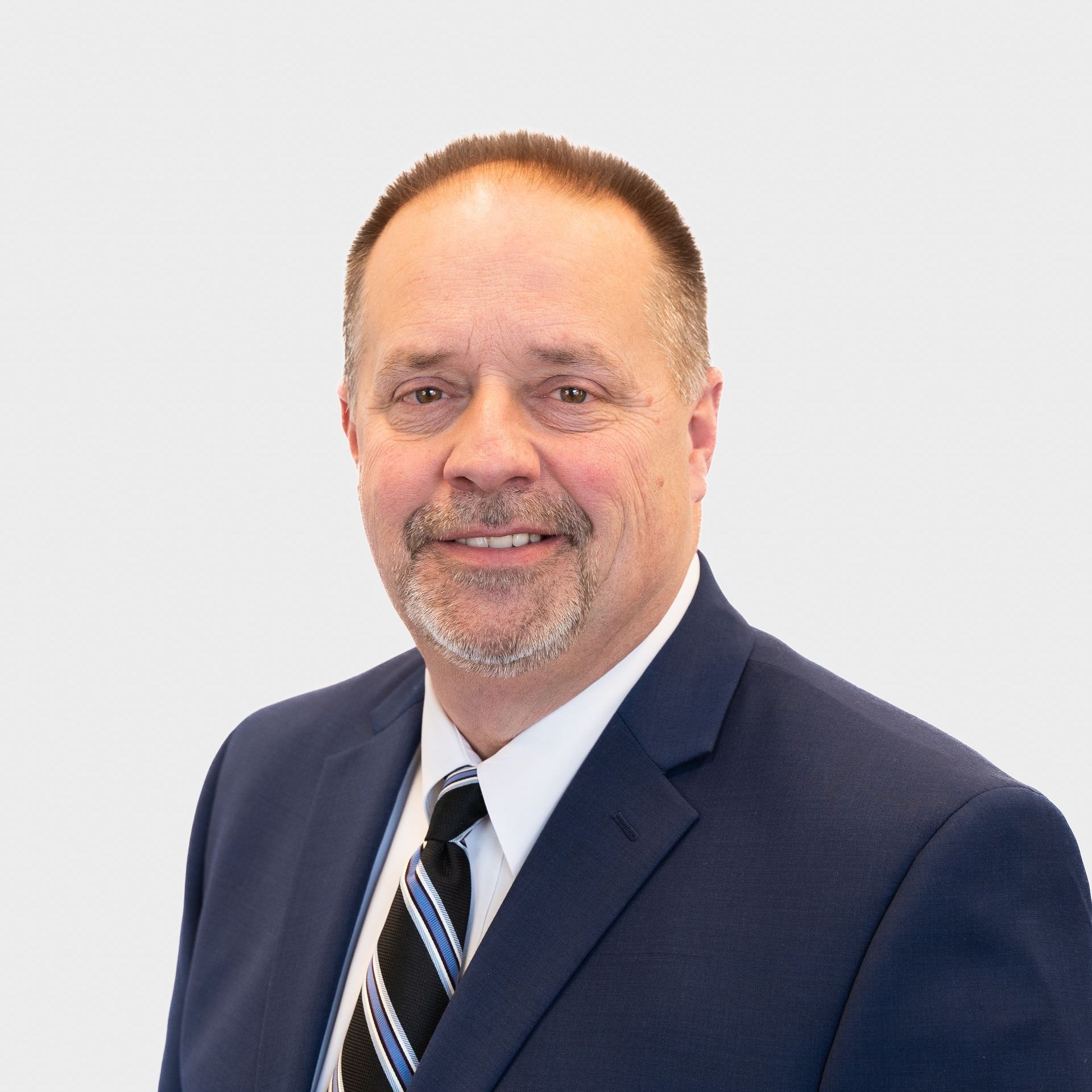 Middle-aged man in navy suit, white shirt, and striped tie smiles against a light gray background.