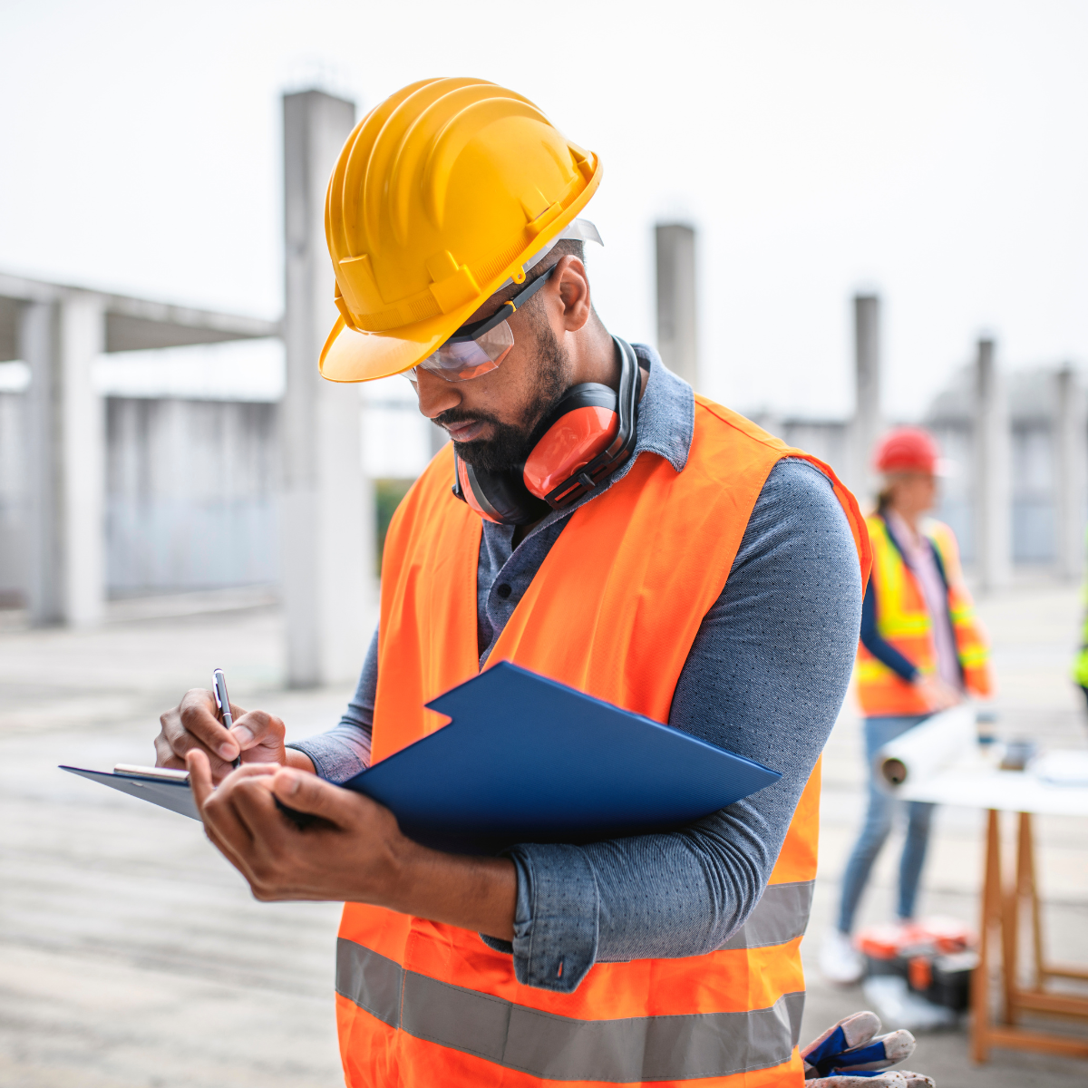 Construction worker in yellow hard hat and orange vest taking notes at a construction site.