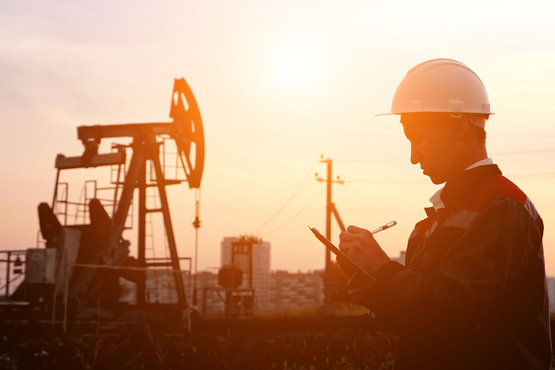 Oil worker in hard hat checking a tablet beside an oil pump at sunset
