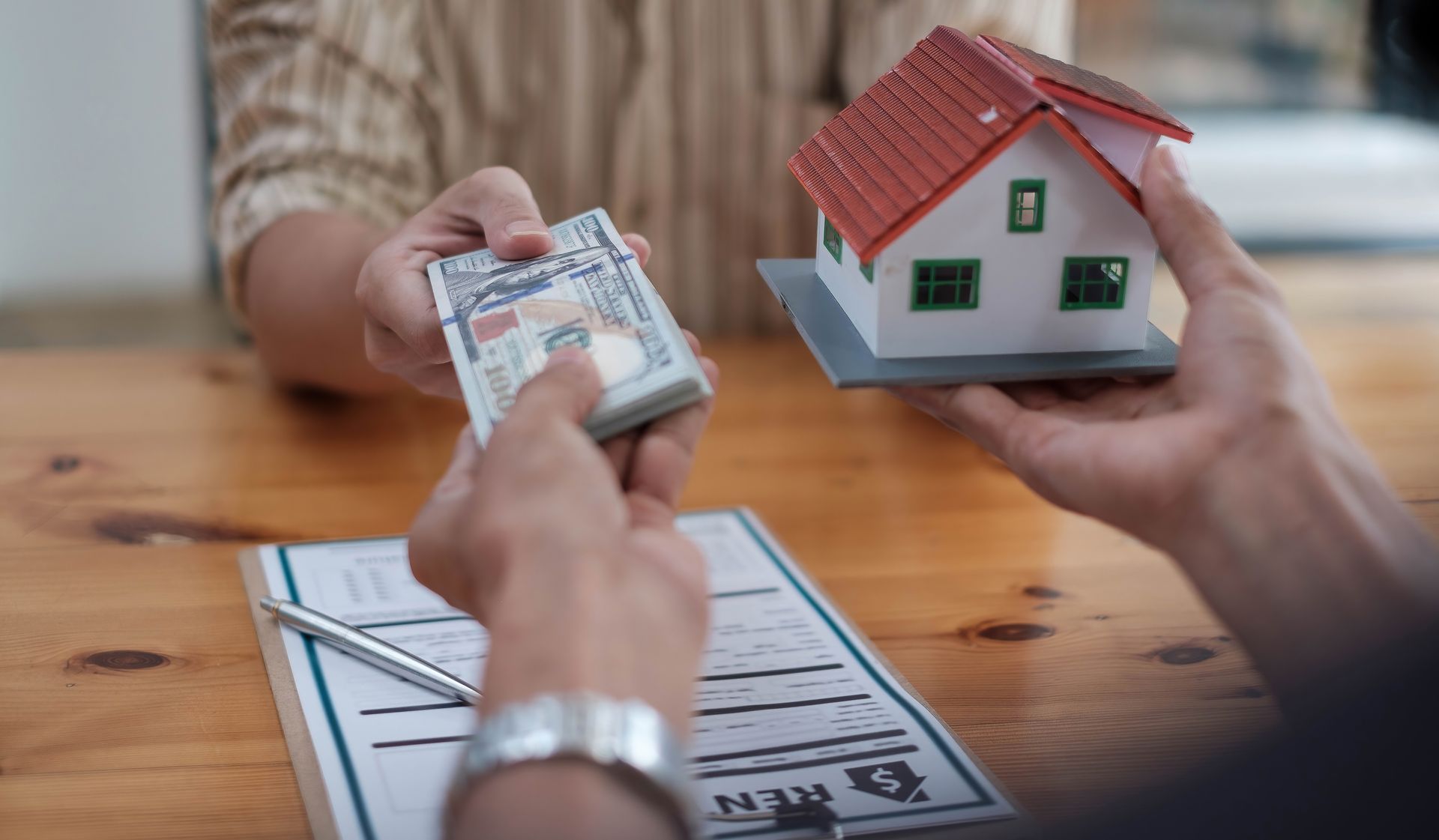 Person hands cash to another holding a miniature house; paperwork and pen on the table.