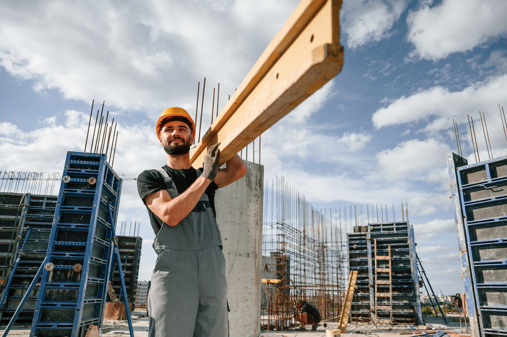 Construction worker carrying a long wooden beam at a building site under a cloudy sky