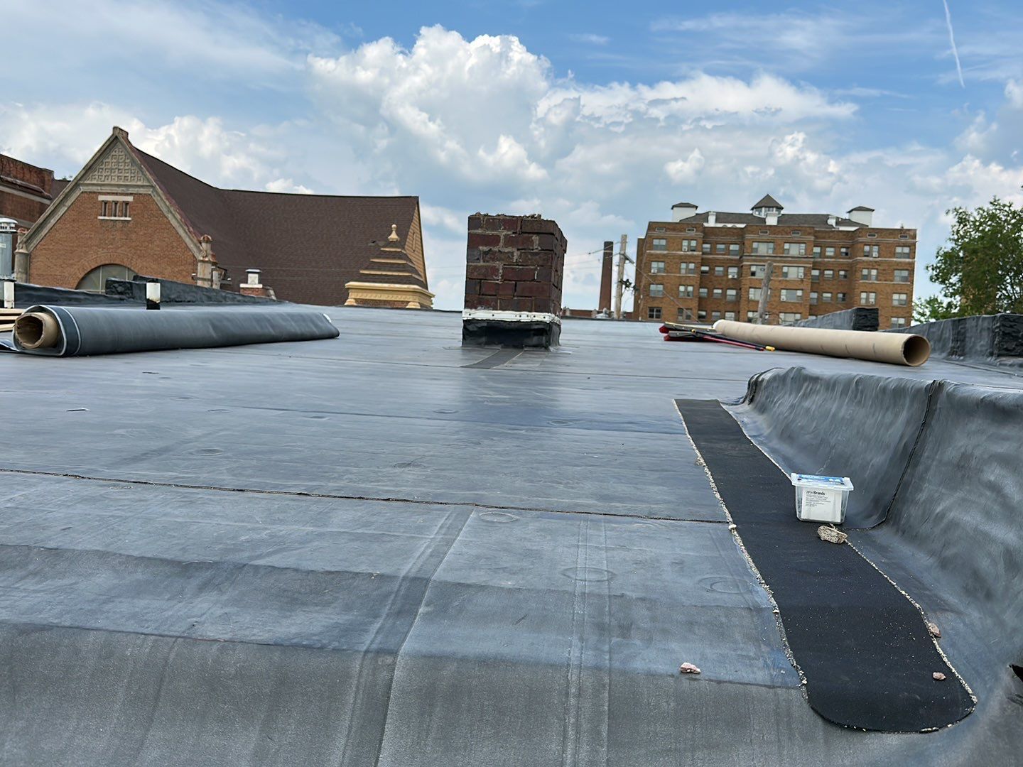 Flat rooftop with black membrane, vents, and nearby brick buildings under a cloudy sky.