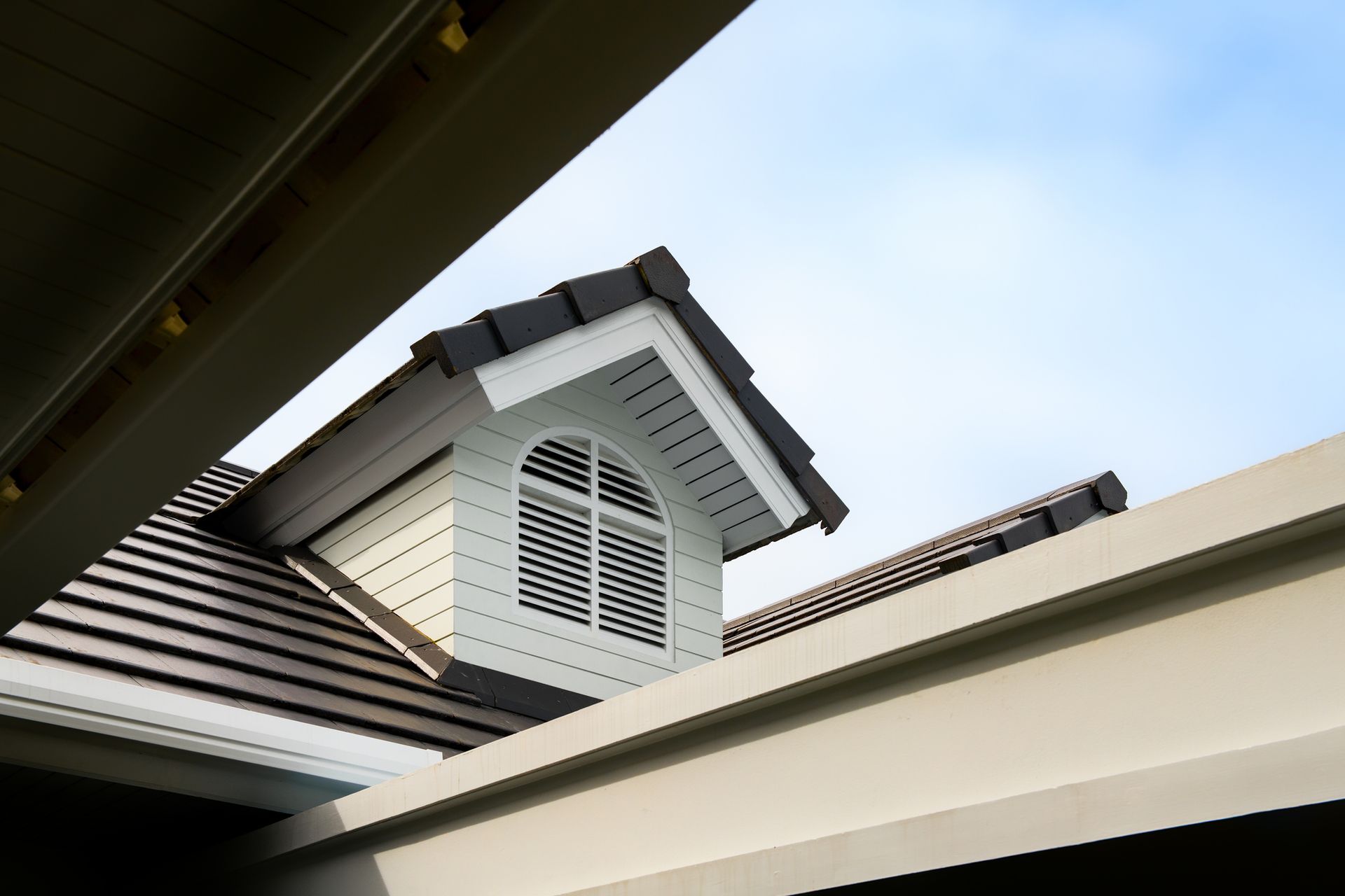 White house dormer with arched shutters and dark roof, seen from below against a blue sky