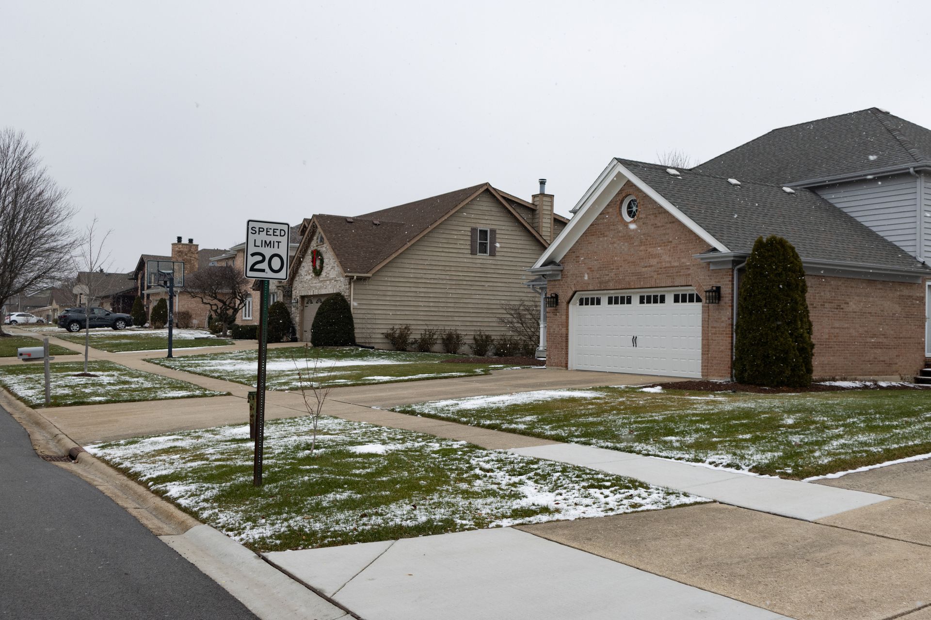 Suburban street with snow-dusted lawns, houses, and a 20 mph speed limit sign.