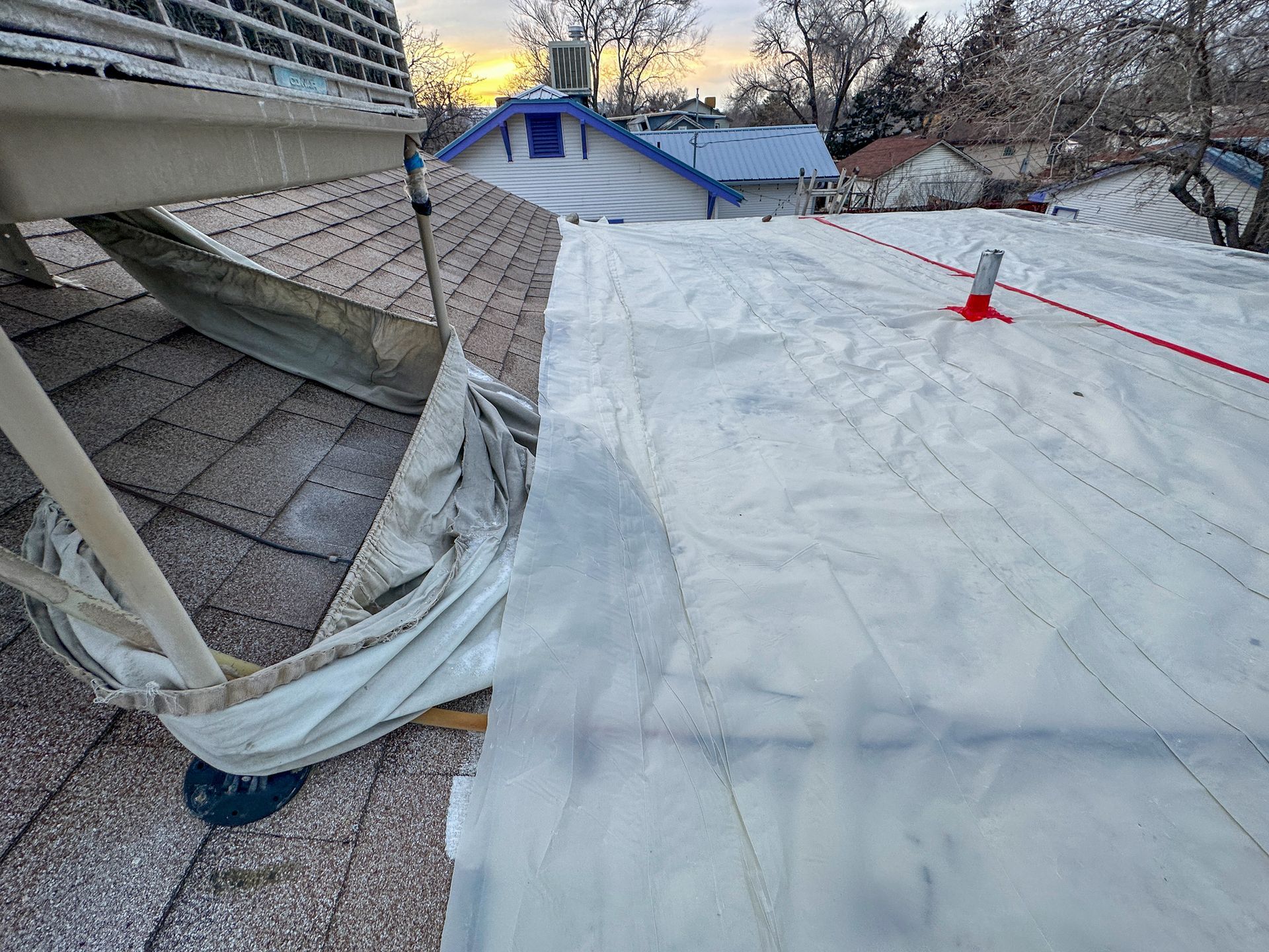Rooftop scene with a large white tarp secured by red rope beside a house and snow-covered yard
