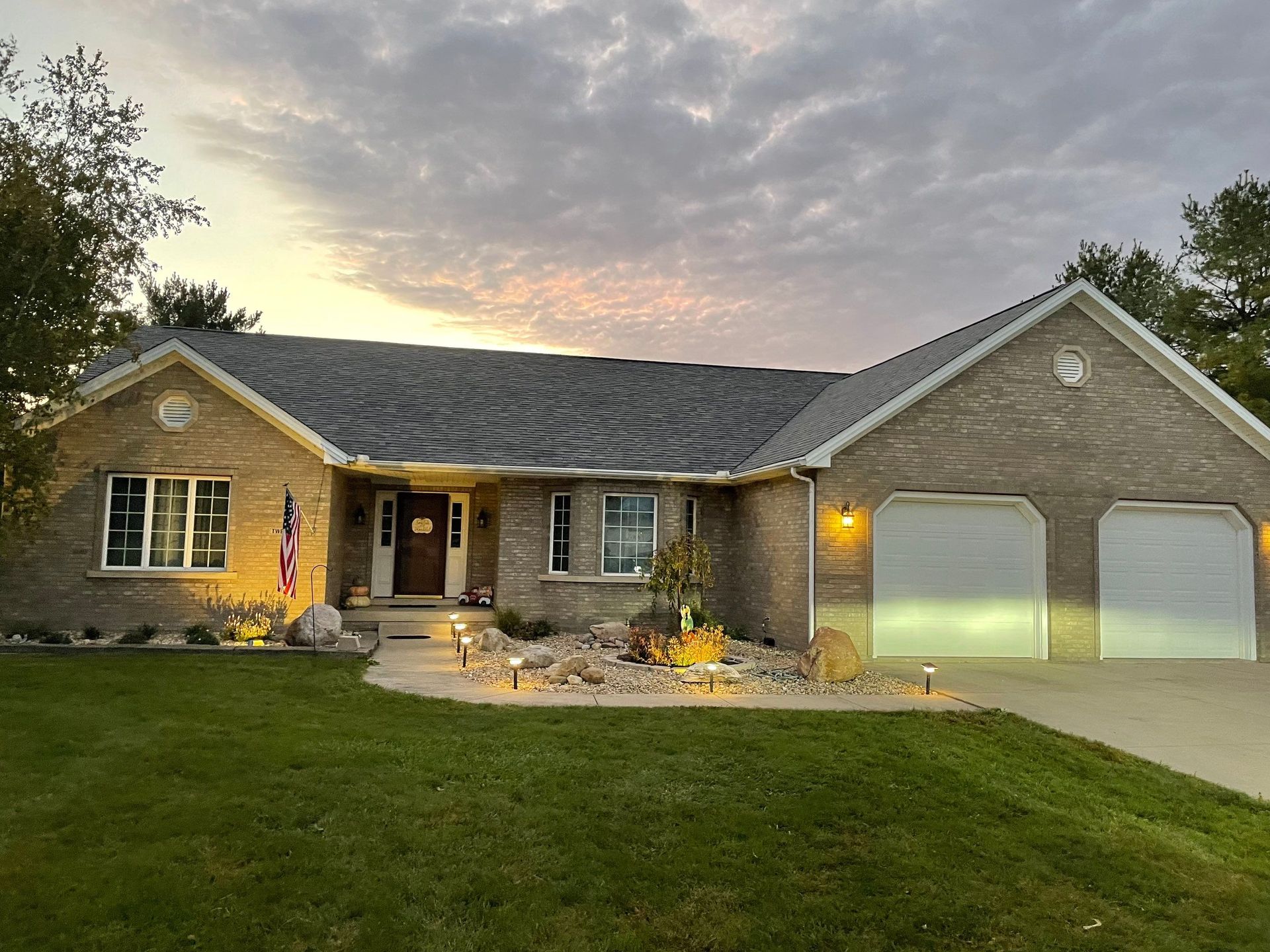 Suburban brick house at dusk with lit porch, two-car garage, and American flag by the front door