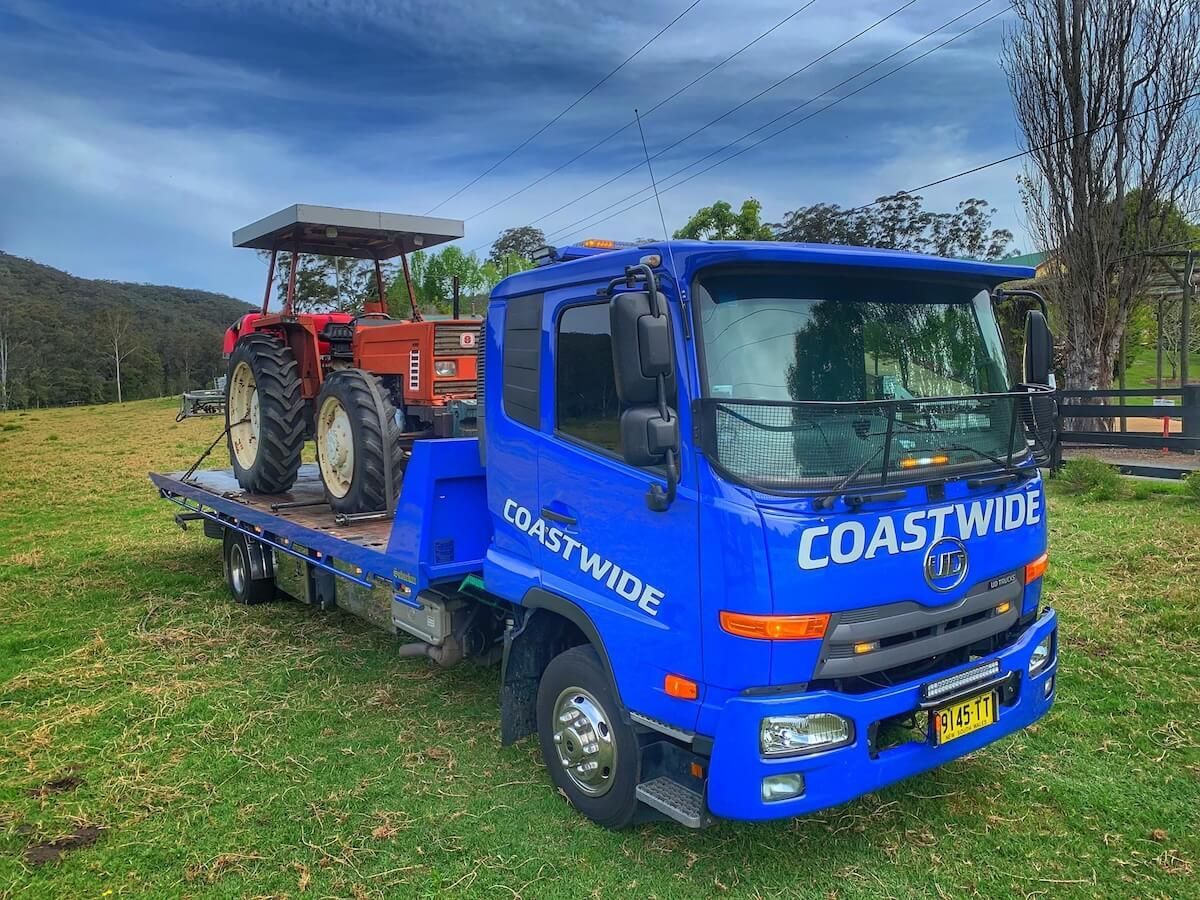 A blue tow truck with a tractor on the back is parked in a grassy field — Coastwide Towing & Transport In Berkeley Vale, NSW