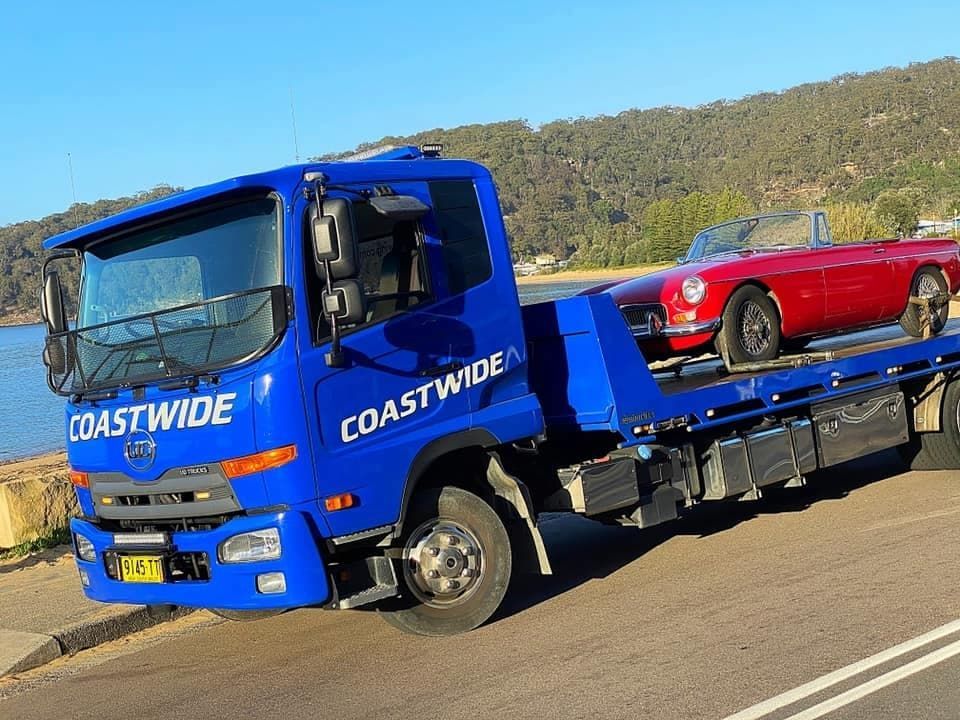 A Blue Tow Truck Labeled Coastwide Carries a Red Vintage Convertible on a Sunny Day