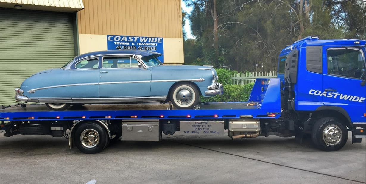 A Blue Tow Truck With A Car On The Back Is Parked In Front Of A Building — Coastwide Towing & Transport In Berkeley Vale, NSW