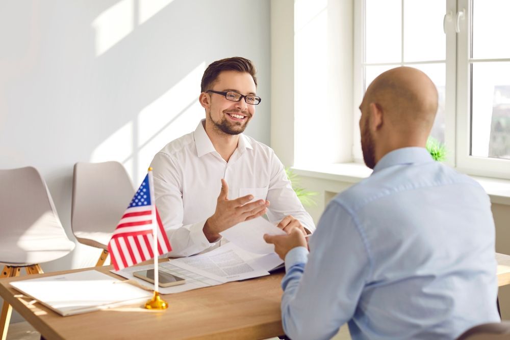 Two men in a bright office discussing across a desk with a small U.S. flag and laptop nearby