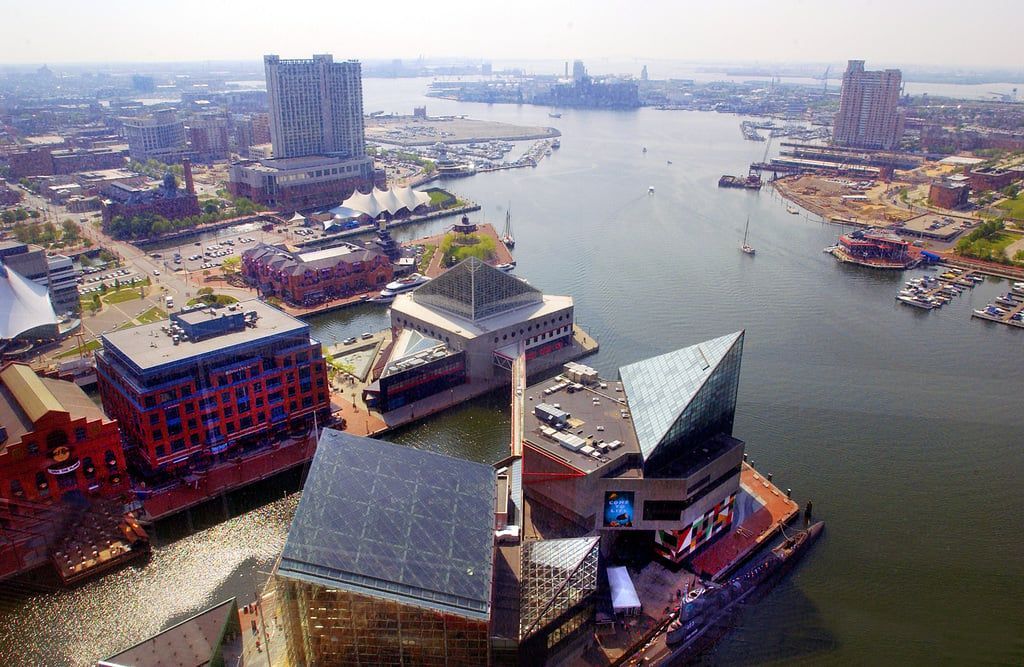 Aerial view of Baltimore's Inner Harbor with buildings, boats, and a wide body of water reflecting the cityscape.