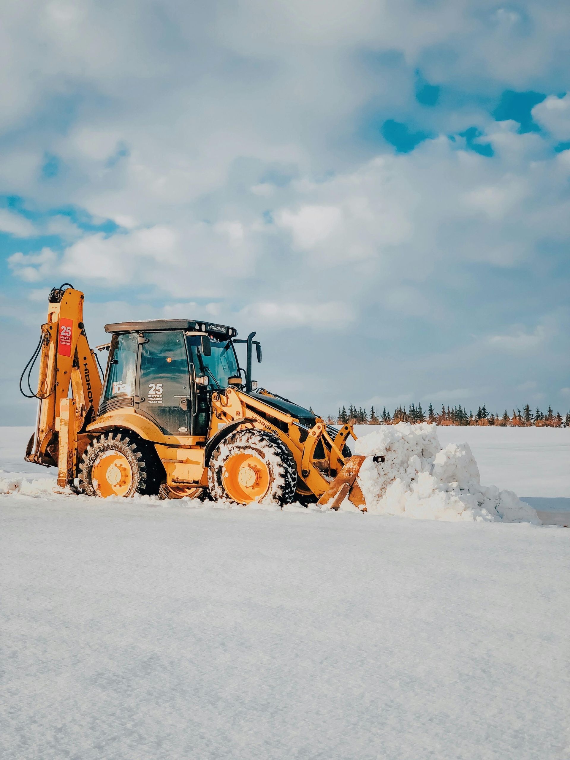 Yellow backhoe plowing snow on a bright, cloudy day.