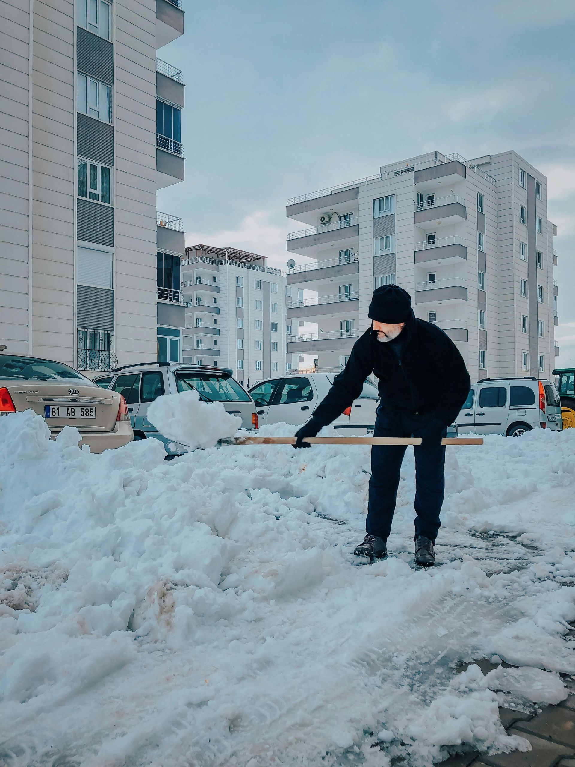 Man shoveling snow in front of apartment buildings on a cloudy day.