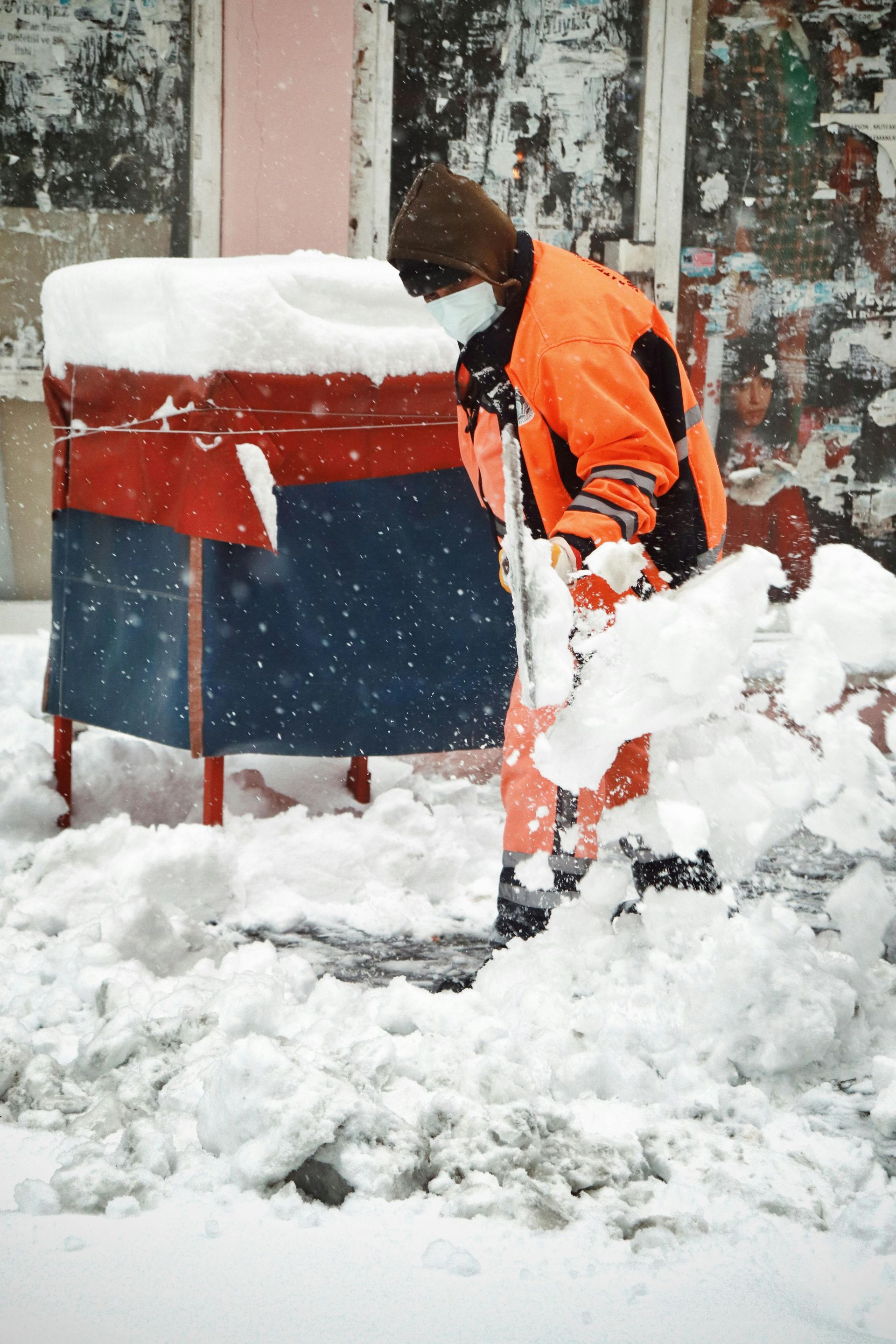 Person in orange jumpsuit shoveling snow near a red and blue stand in a snowy environment.