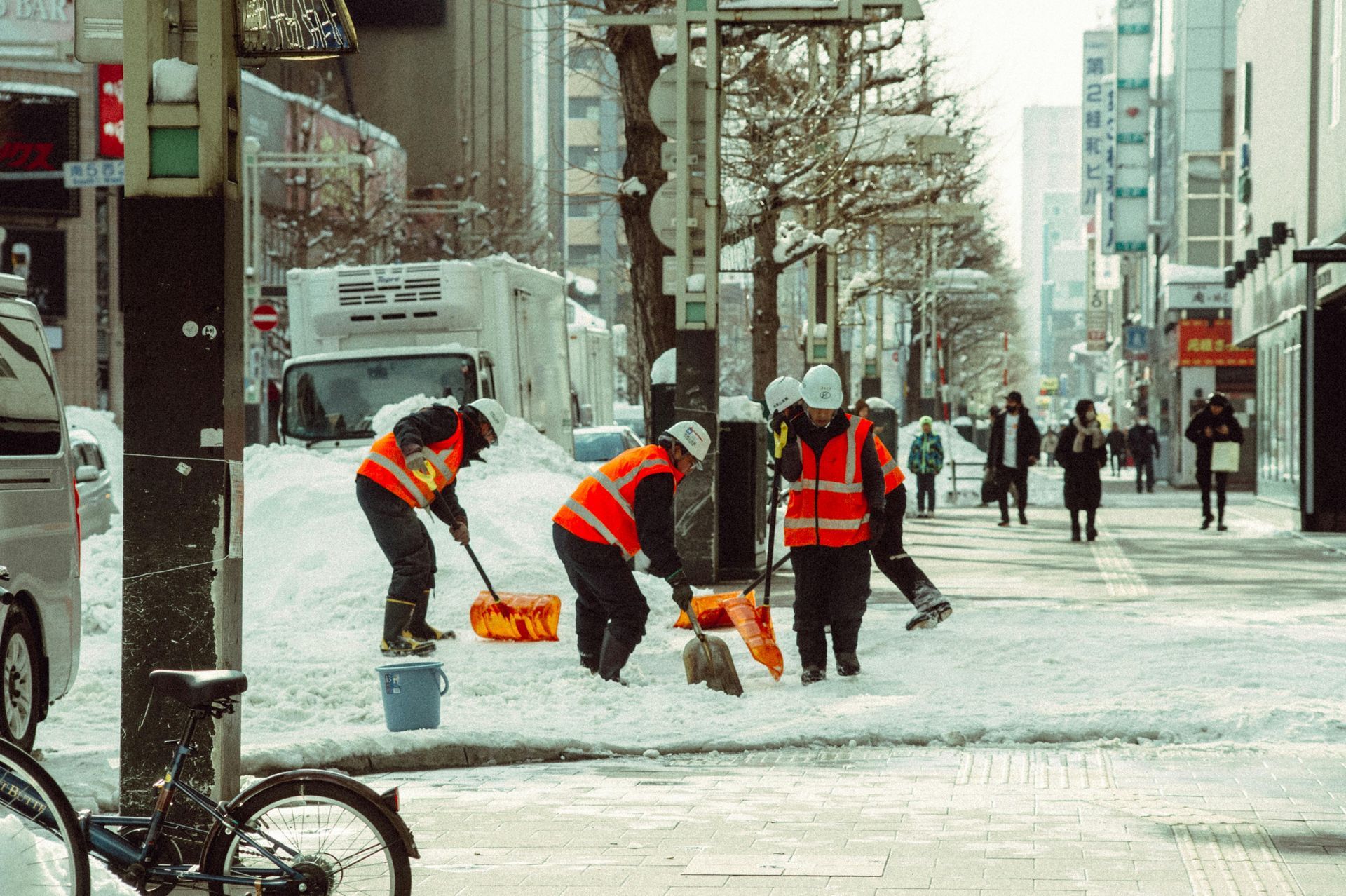 Workers in orange vests shoveling snow on a city sidewalk; buildings and cars in background.