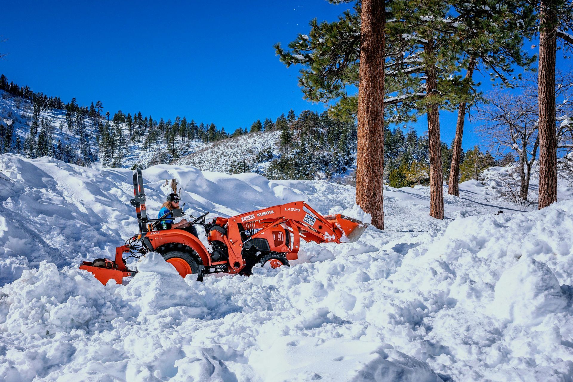 Orange tractor plowing snow on a mountain road, under a blue sky, near pine trees.