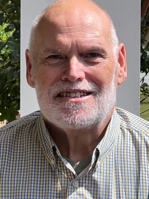 Man with a white beard and bald head wearing a button-down shirt, smiling in front of a white wall.