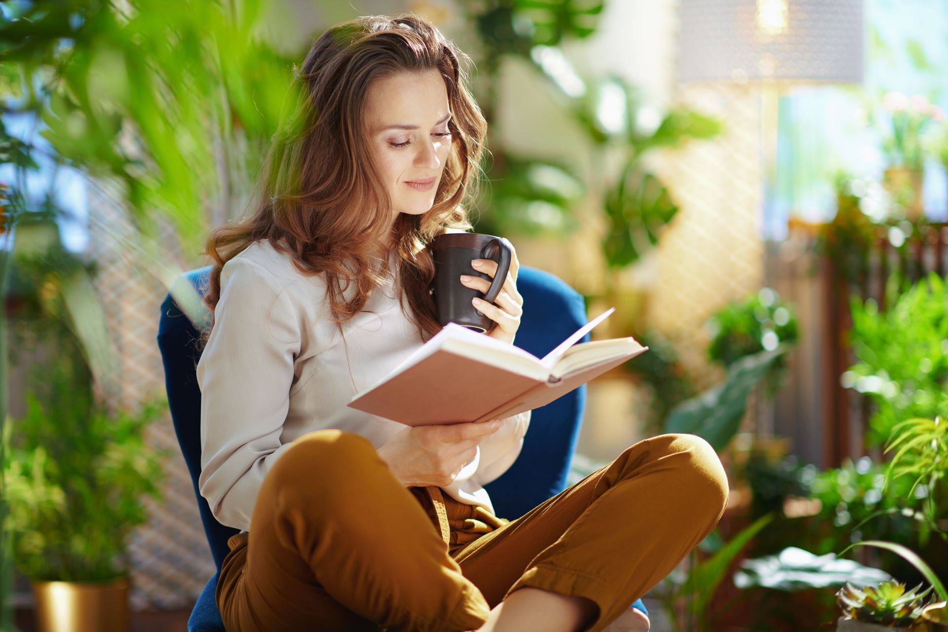 Woman sitting, reading a book, and holding a mug, surrounded by plants and sunlight.