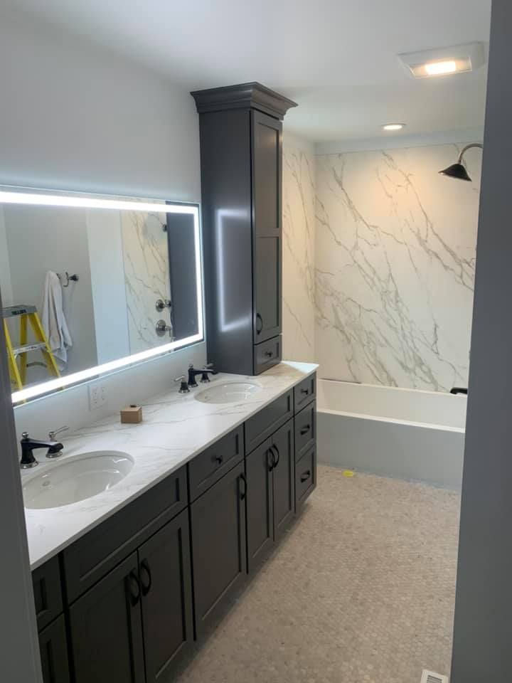 Modern bathroom with gray vanity cabinets, marble-look countertops and tub surround, and an illuminated mirror.