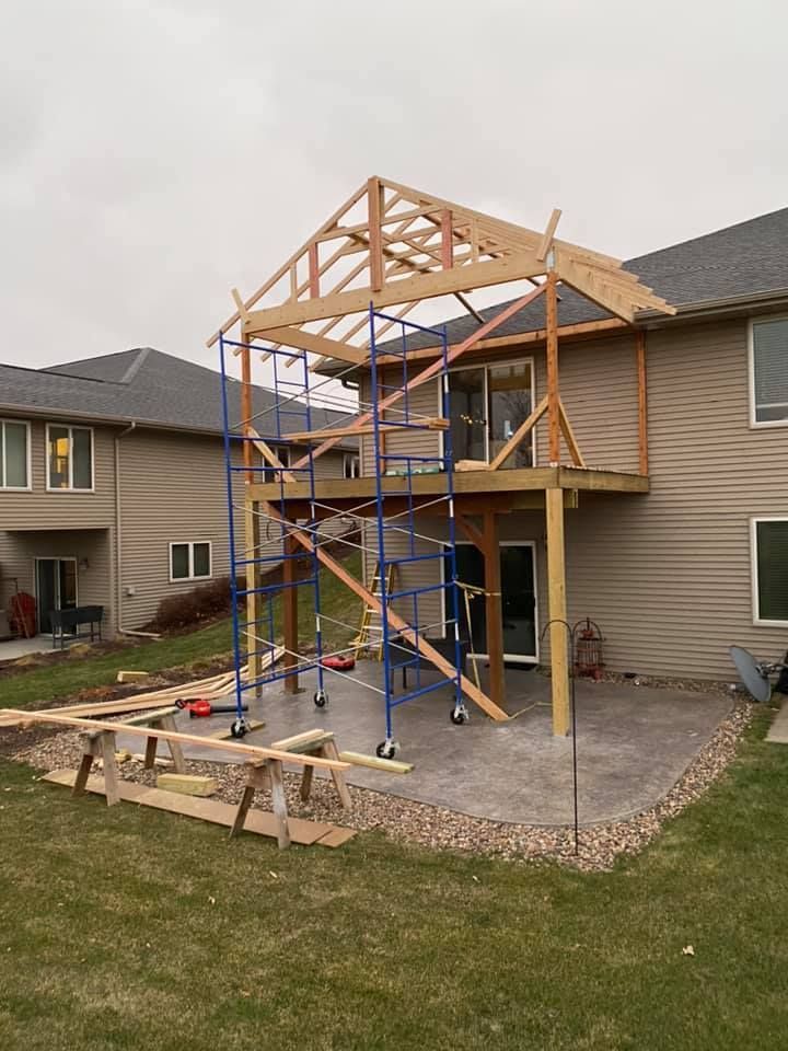 A partially framed wooden porch addition under construction on the back of a tan suburban home with a concrete patio.
