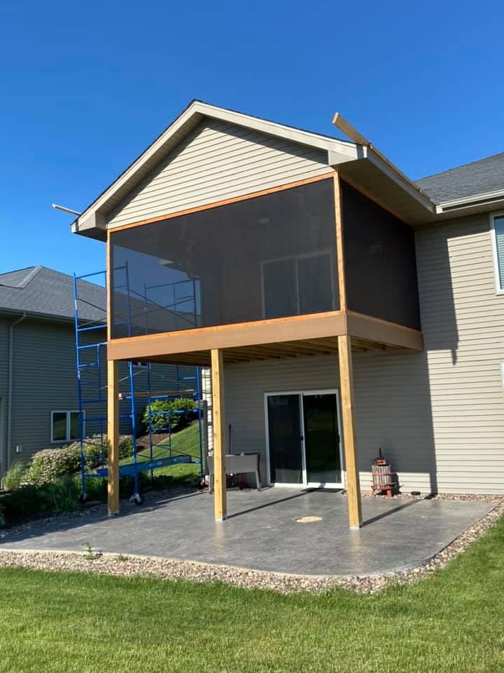 A newly constructed second-story screened-in porch with light siding stands above a gray patio on a sunny day.