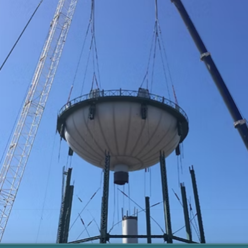 Black water tank atop rusty metal tower against a blue sky with clouds.
