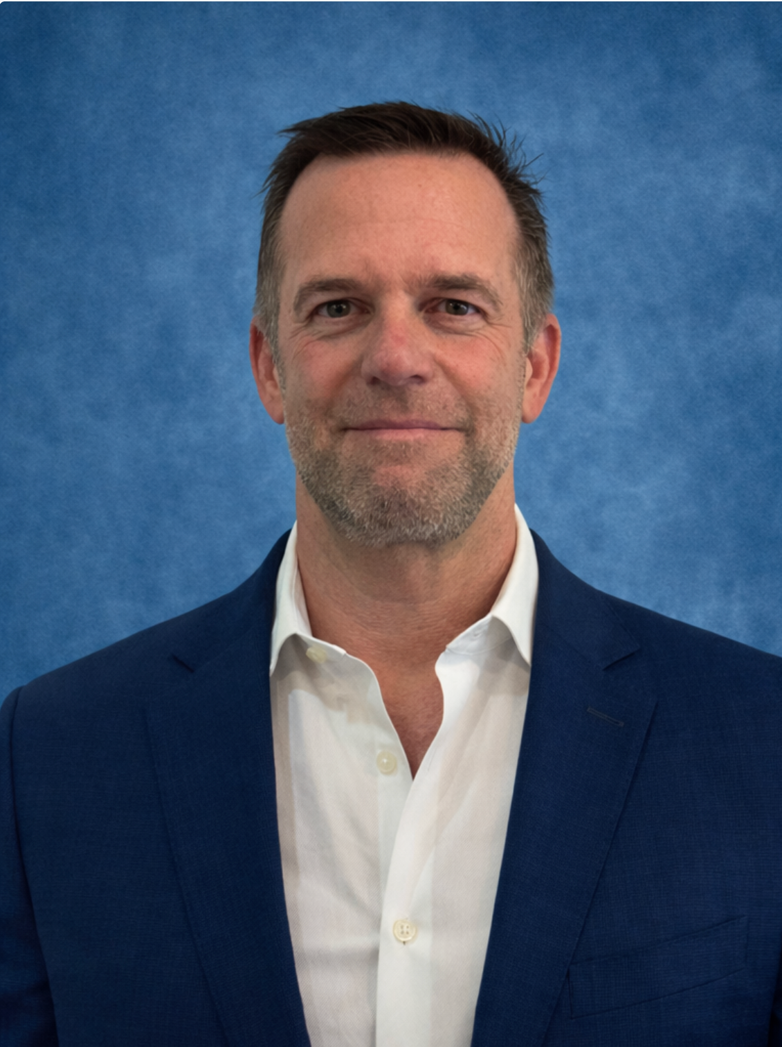 Man in blue blazer and white shirt smiles, blue background.