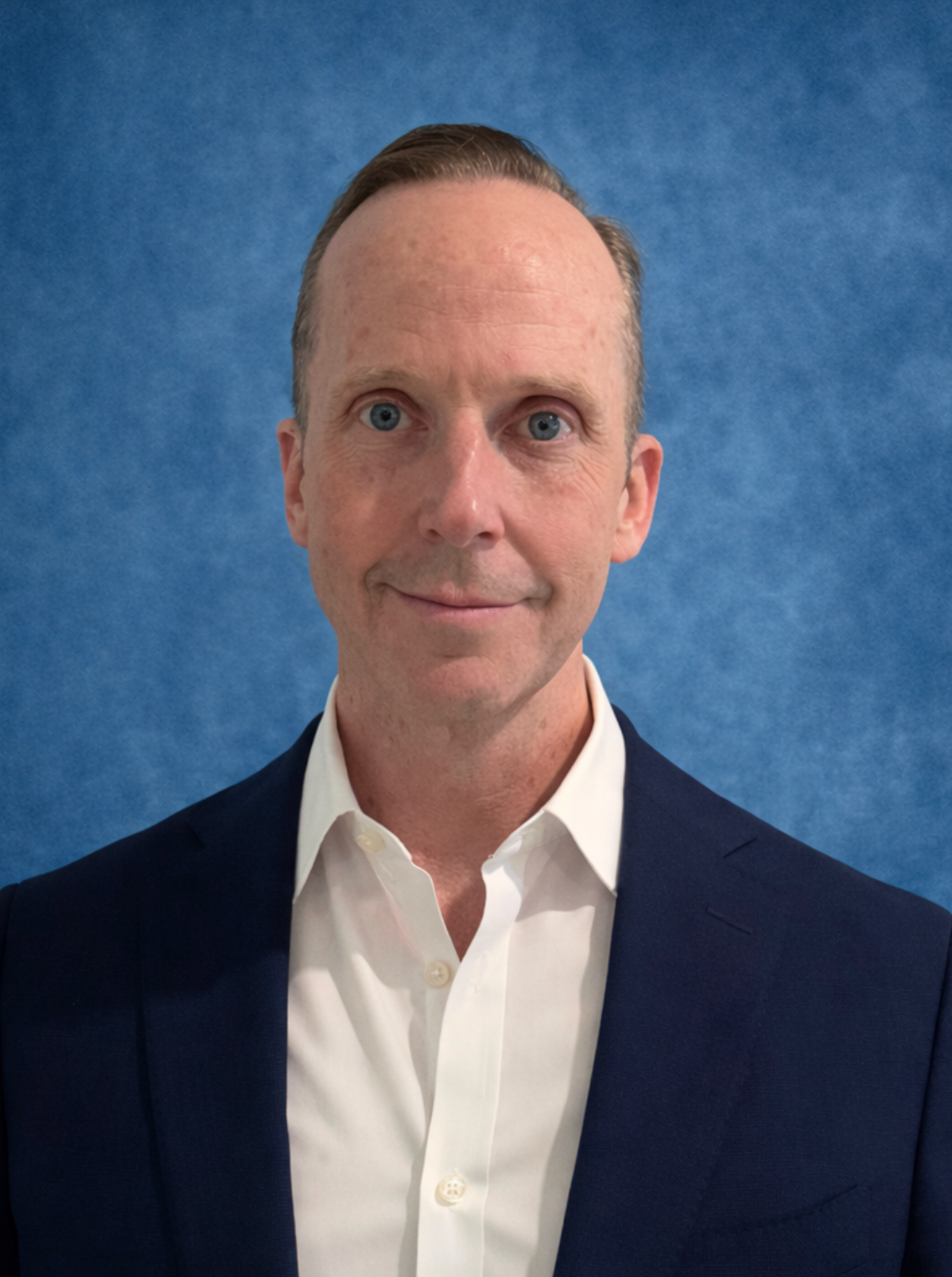 Man in a navy blazer and white shirt against a blue backdrop; looking forward.