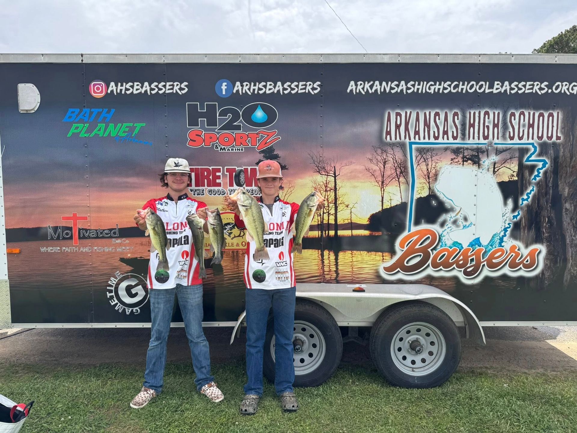 Two people in matching jerseys stand in front of a Bassmaster fishing trailer at an outdoor event.