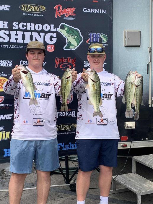 Two anglers in white jerseys hold bass on a tournament stage with sponsor backdrop