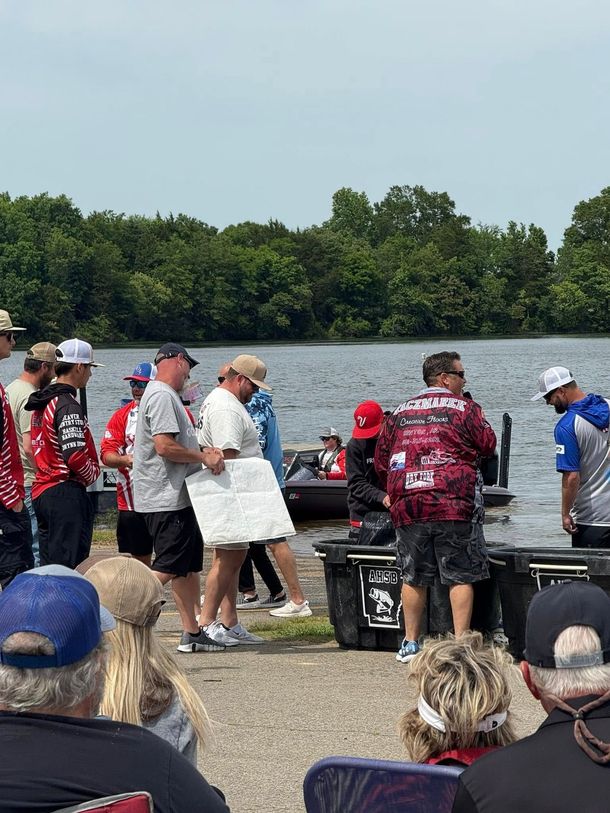 People gathered outdoors near tables by the water, with a small crowd in sports jerseys and hats.
