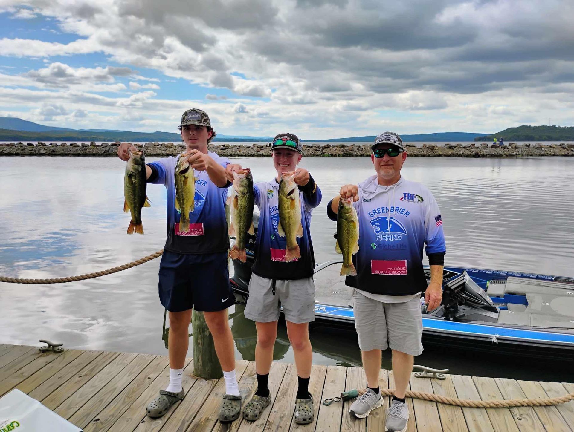 Three anglers stand on a dock holding large fish, with a calm harbor and cloudy sky behind them.