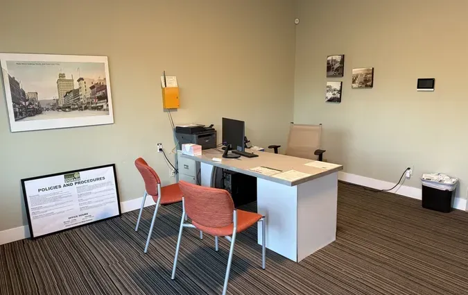 A modest office with a desk, computer, two orange chairs, a large framed print on the wall, and a document on the floor.