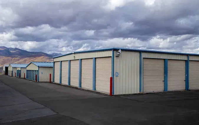 A row of beige storage units with rolling doors under a cloudy sky, with mountains visible in the distance.