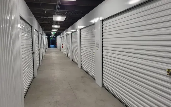 A long, narrow indoor hallway lined with white roll-up metal doors for storage units under fluorescent lighting.