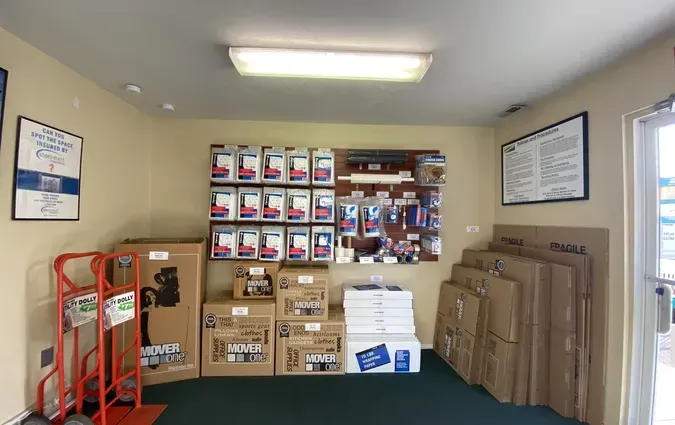 A storage supply shop interior with boxes, moving equipment, and merchandise displayed on wall pegs and cardboard displays.