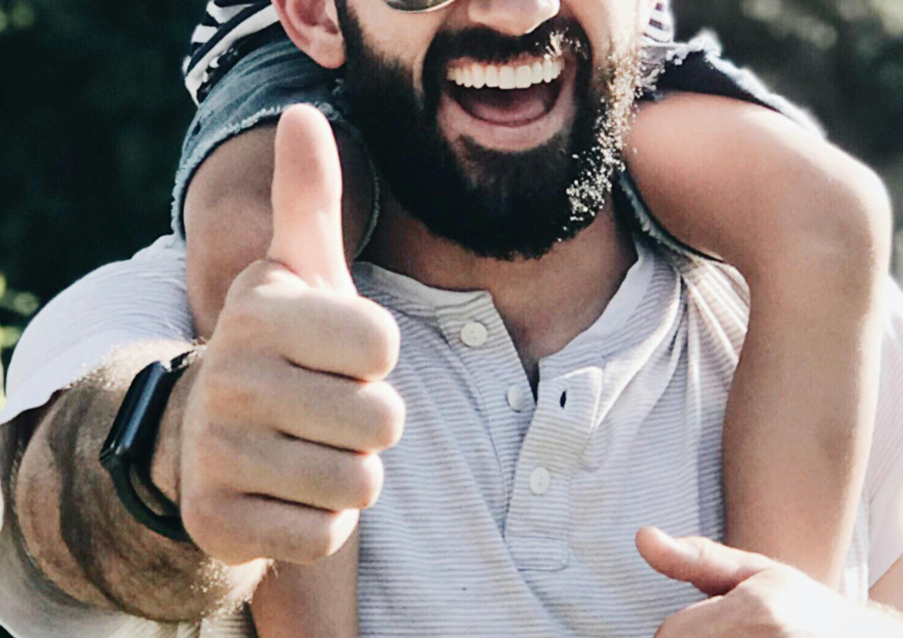 A man is carrying a child on his shoulders and giving a thumbs up.