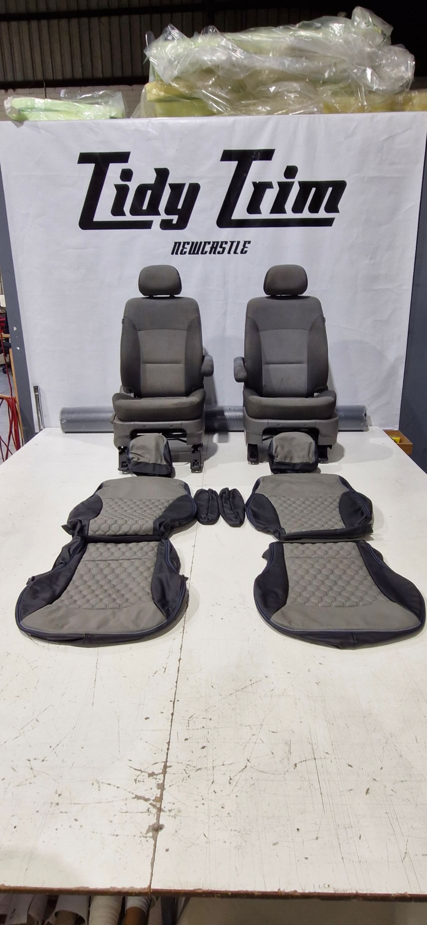 Two automotive seats and matching gray and black patterned upholstery covers displayed on a white table under a sign — Tidy Trim Newcastle in Carrington, NSW