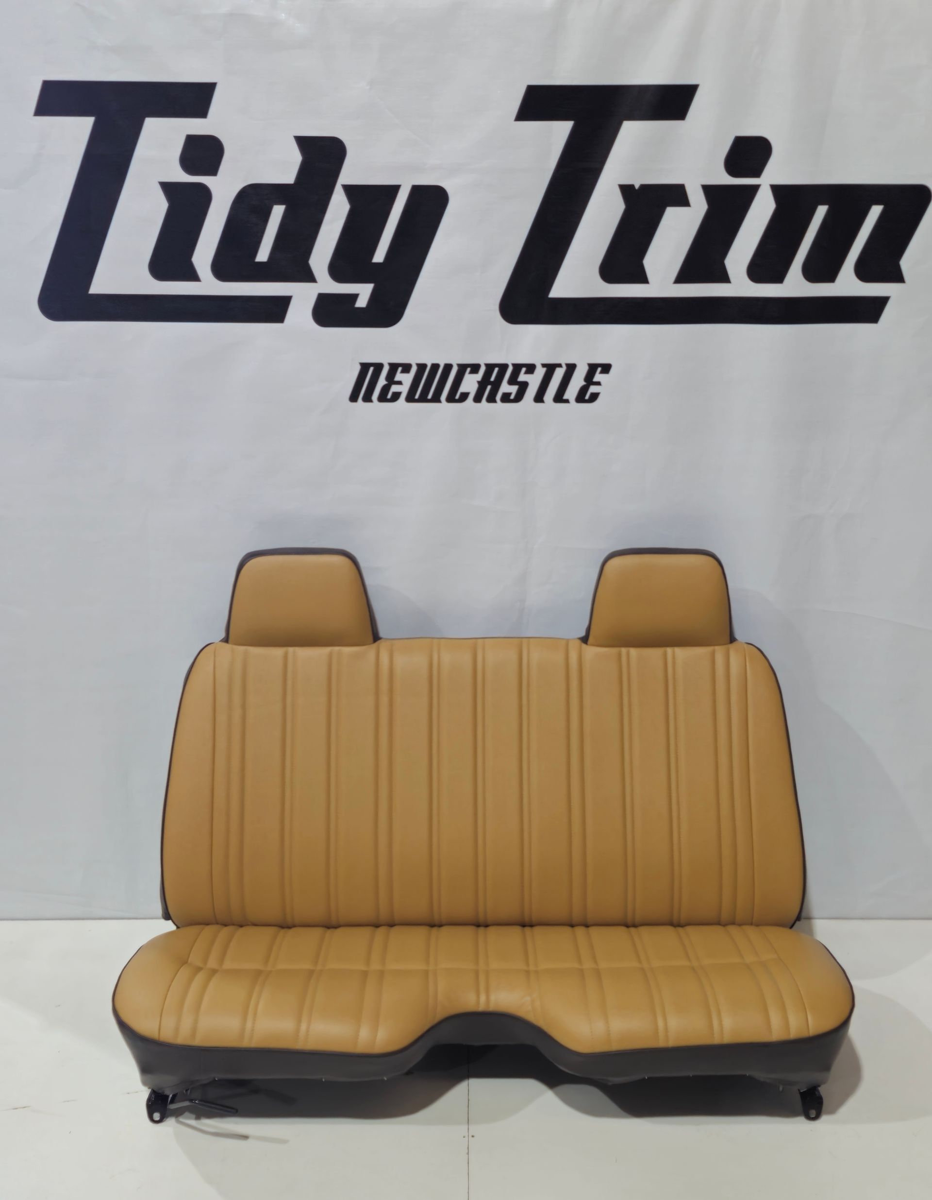 Tan bench seat with vertical stitching and two headrests, set against a Tidy Trim Newcastle branded backdrop  — Tidy Trim Newcastle in Carrington, NSW