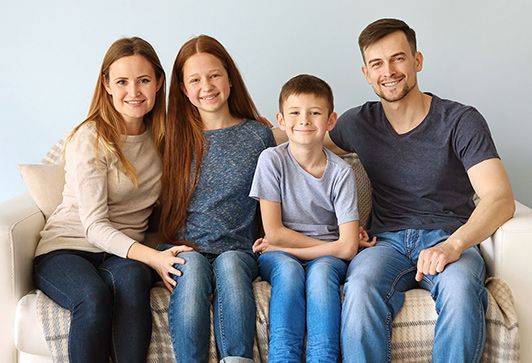 Family of four, smiling, sitting on a couch together, blue wall background.