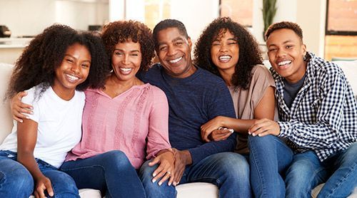 Family of five smiling and sitting on a couch; indoor setting, mixed skin tones.